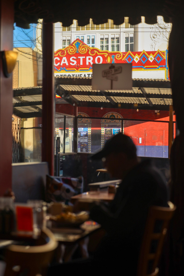 A person sits in a dimly lit cafe with a view of the colorful Castro Theatre sign outside, framed by windows and a street awning.